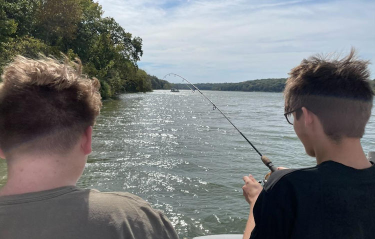 two young men sitting on a boat in the middle of a lake fishing pictured from behind