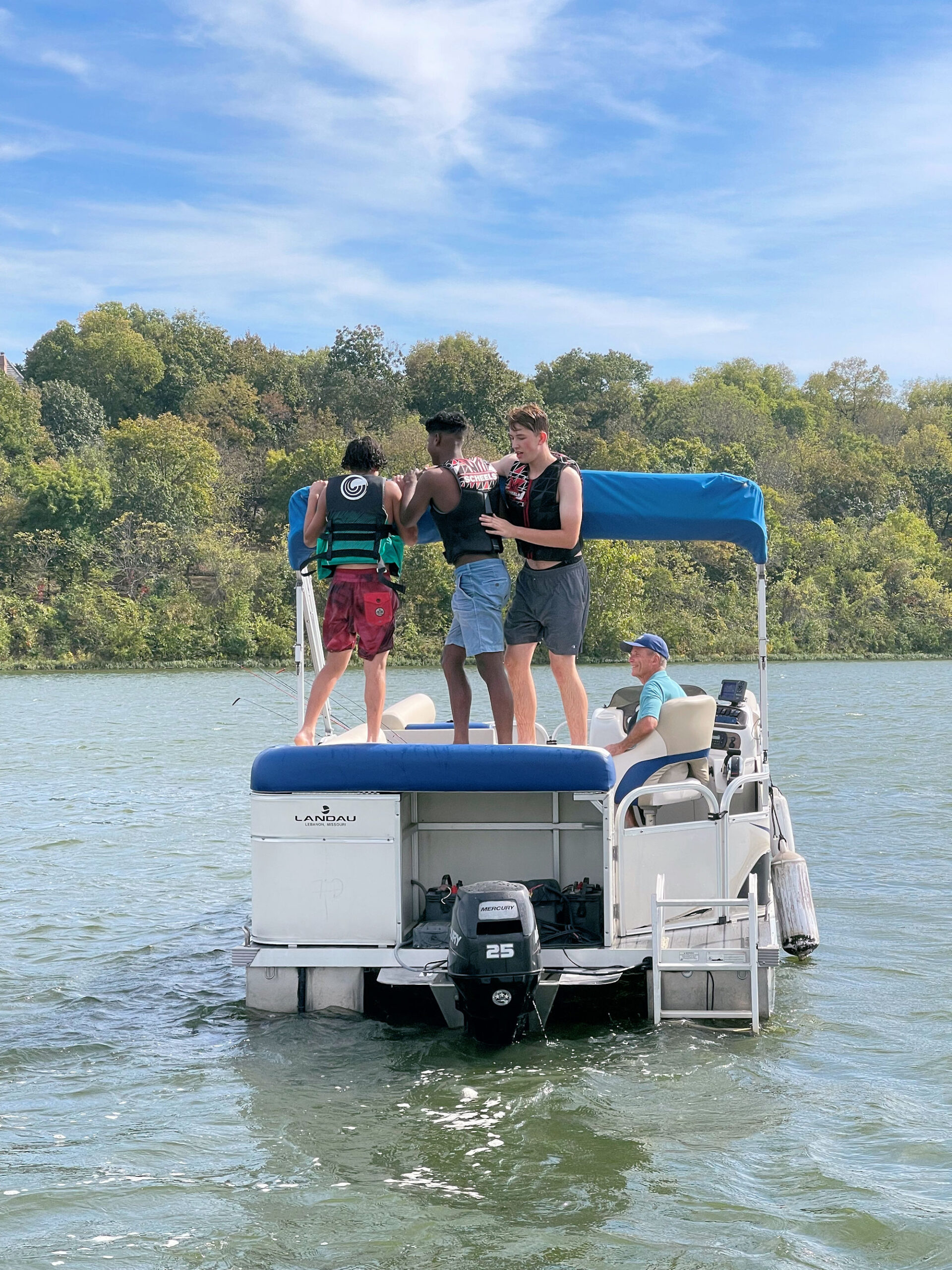 three young men standing on a white boat wearing lifejackets in the middle of a blue lake with green trees in the background